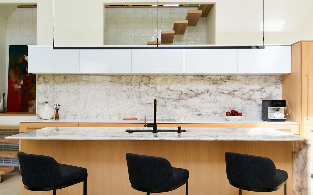 Bright, modern kitchen with marble countertops and backsplash, light wood cabinetry, and three dark gray bar stools at the island. Floating stairs visible in background. Mountain Contemporary design.