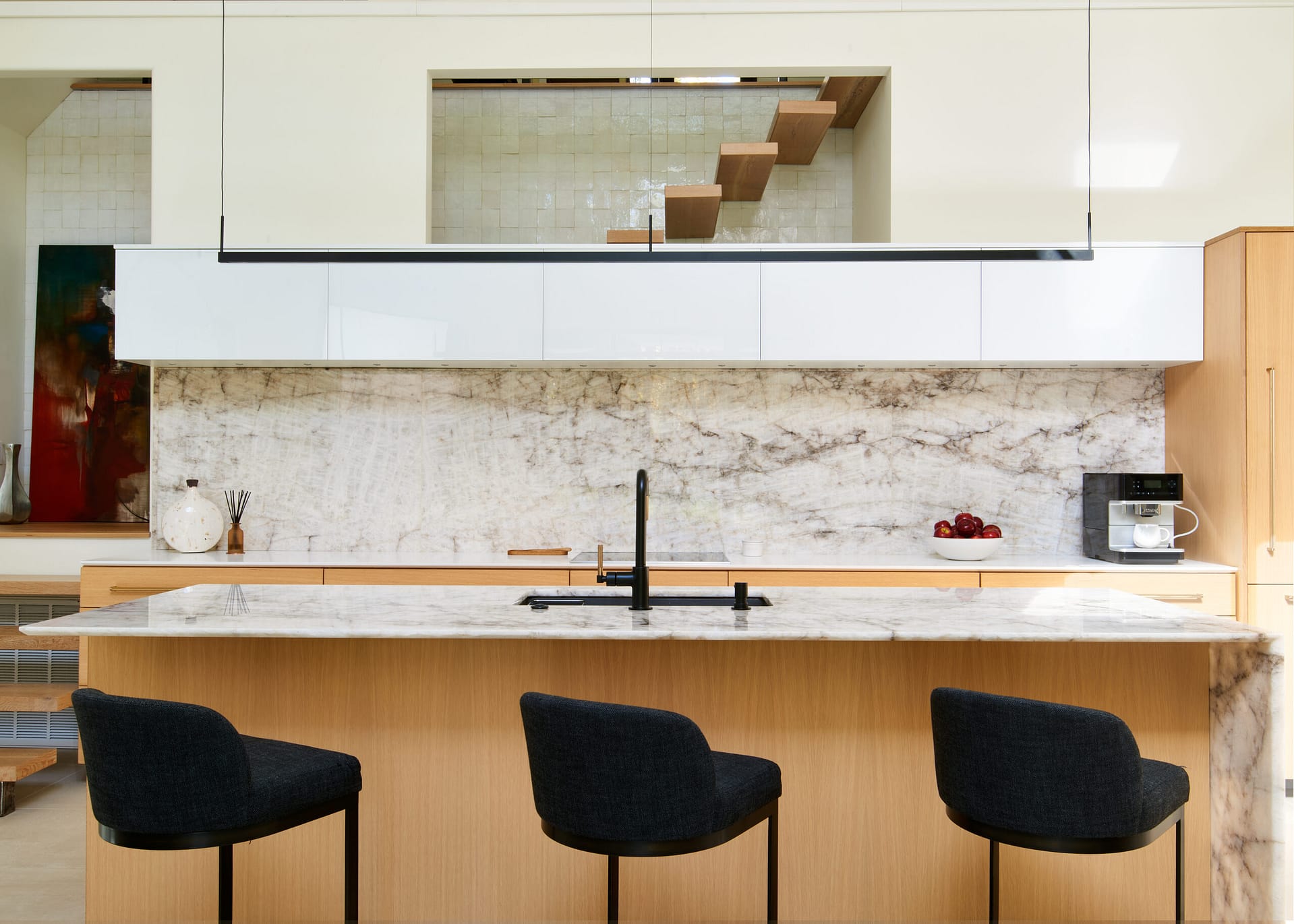 Bright, modern kitchen with marble countertops and backsplash, light wood cabinetry, and three dark gray bar stools at the island. Floating stairs visible in background. Mountain Contemporary design.